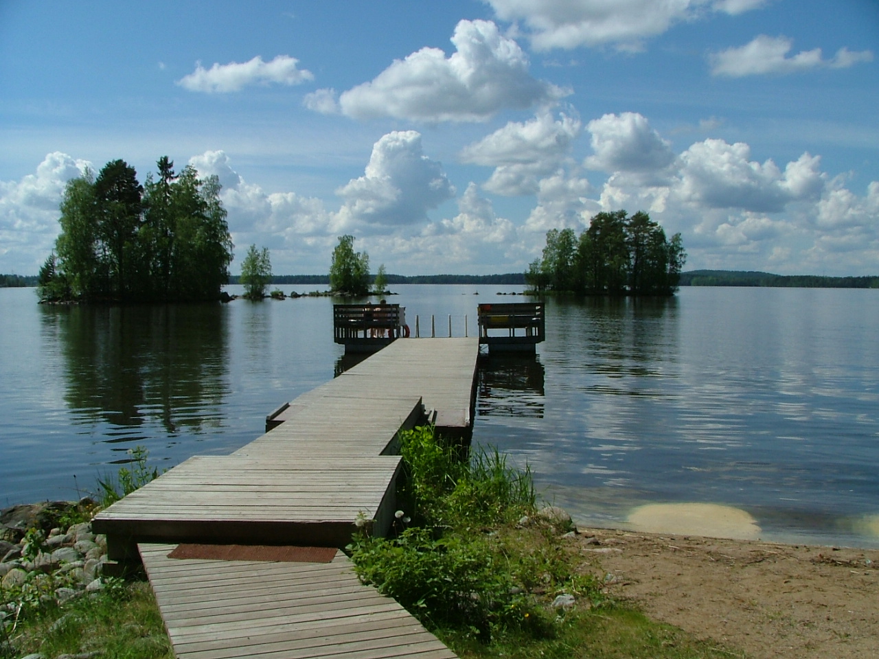 Kuvassa on pitkä laituri ja näkymä järvelle päin. Laiturin päässä olevalla penkillä istuu kaksi uimaria. Järvessä näkyy kaksi pientä saarta. Ilma on aurinkoinen. Taivaalla muutamia kumpupilviä.