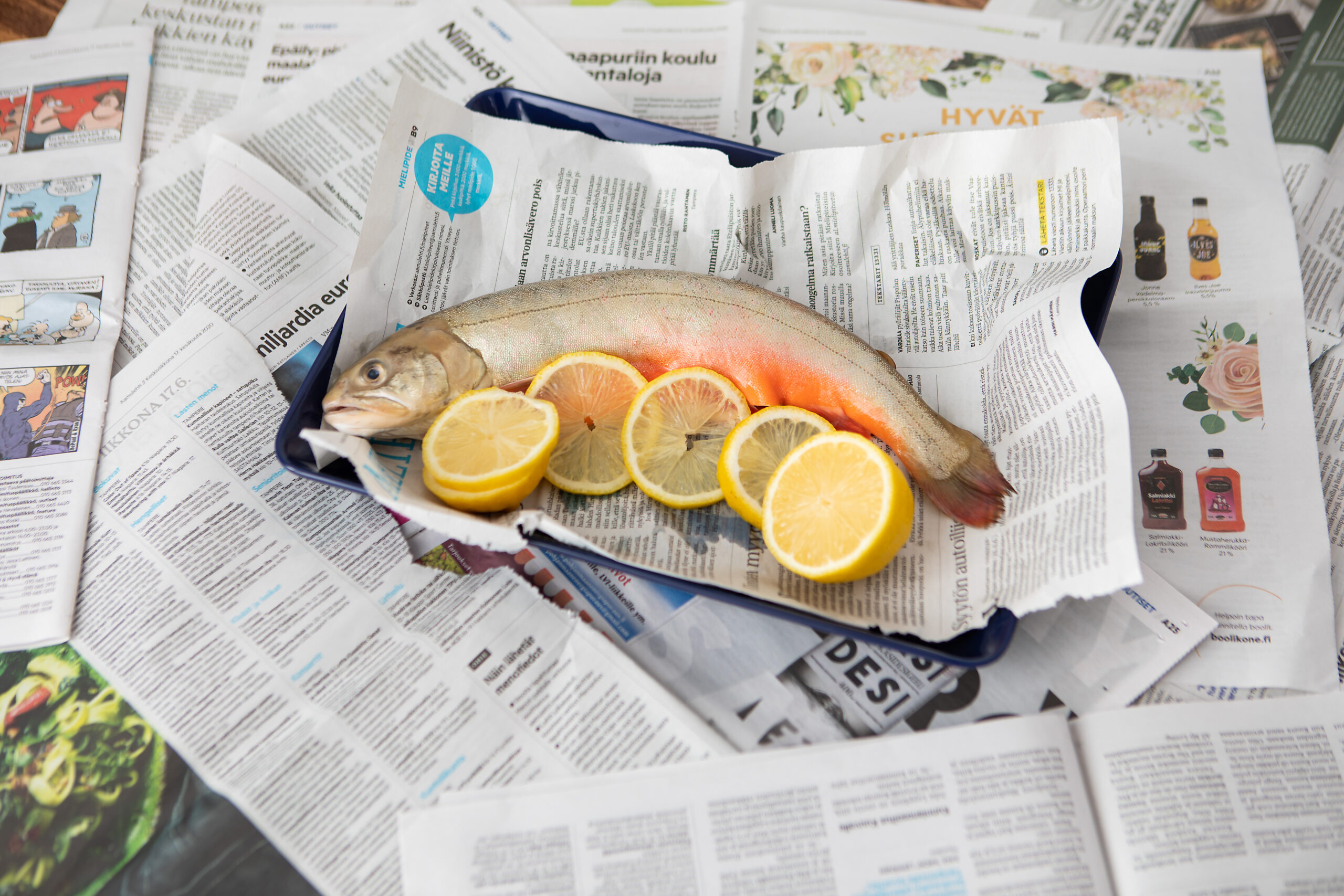 Kuvassa sinisellä lautasella sanomalehtipaperin päällä kala, vieressä sitruunan lohkoja. Lautasen alla pöydällä sanomalehtiä avattuna.