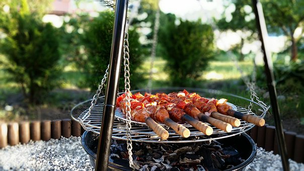 Kuvassa grilli, jossa paistumassa grillivartaita, joissa on lihaa ja tomaatteja. Taustalla näkyy aurinkoinen kesäinen luonto.