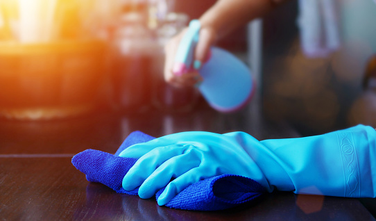 Kuvassa käsi, jossa sininen siivoushansikas, pyyhkiin kirkkaan sinisellä siivousrätillä pöytää. Taustalla näkyy  käsi, jossa vaaleansininen suihkepullo.